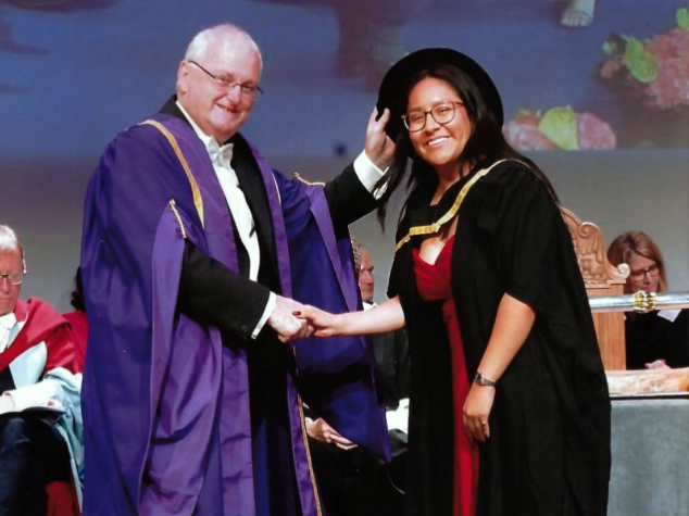 A photo of a woman wearing graduation regalia receiving a diploma
