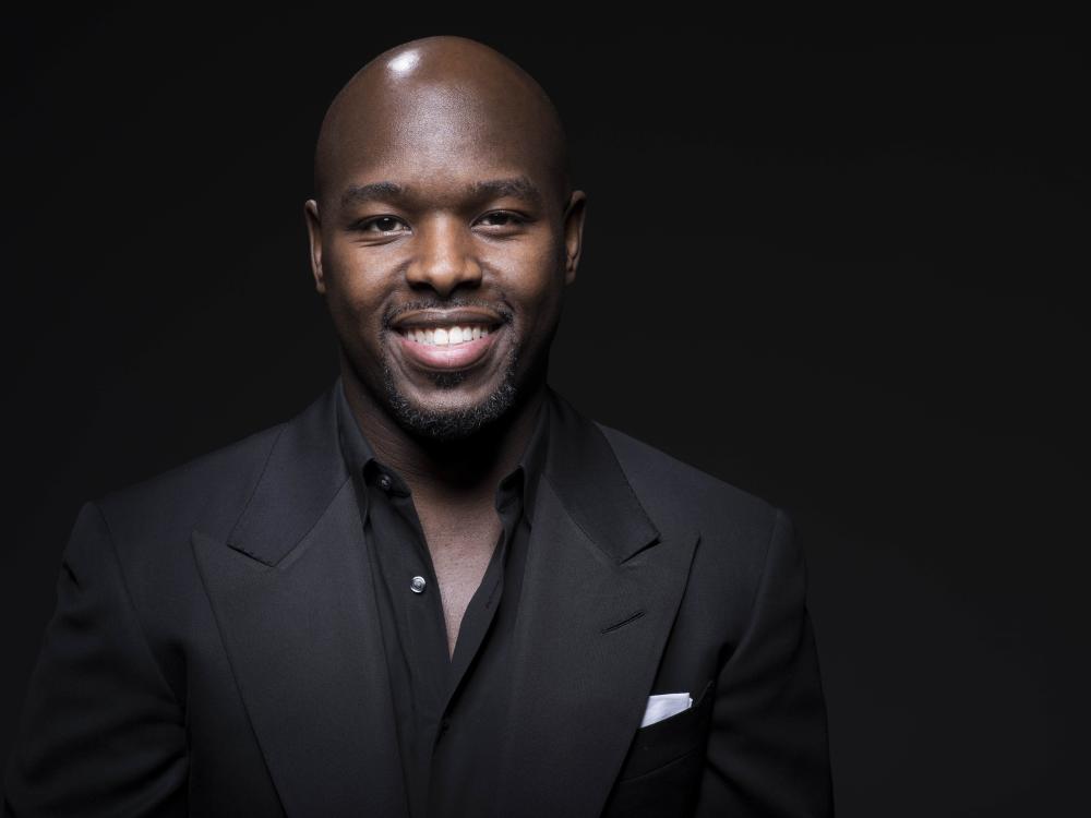 A smiling Black man wearing a dark suit stands against a dark wall.