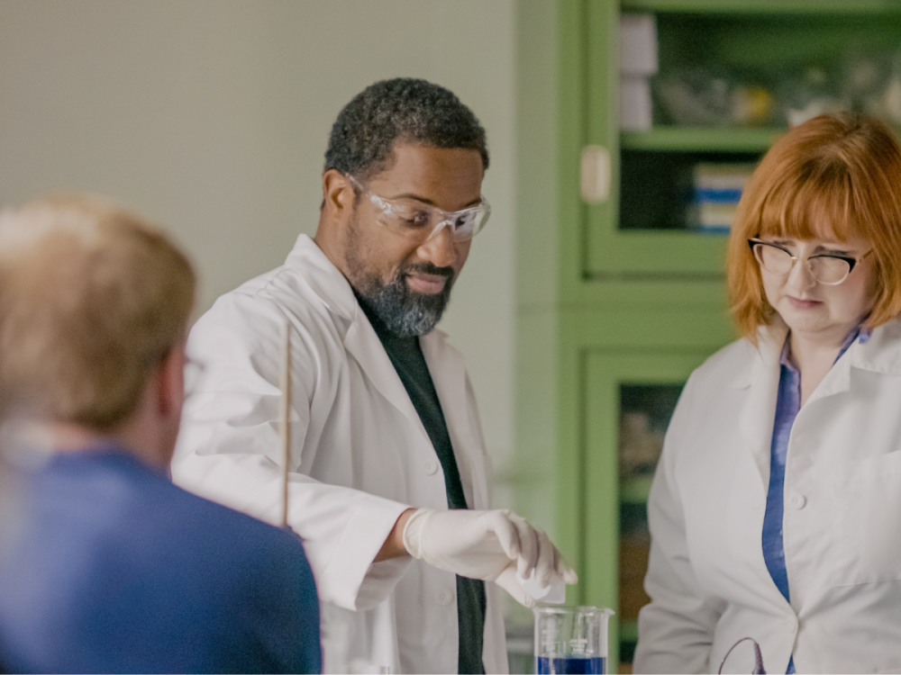 Two individuals in lab coats pouring a liquid into a beaker