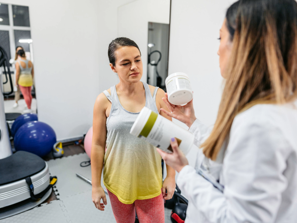 A nutritionist holds supplements while talking to a woman in a gym.