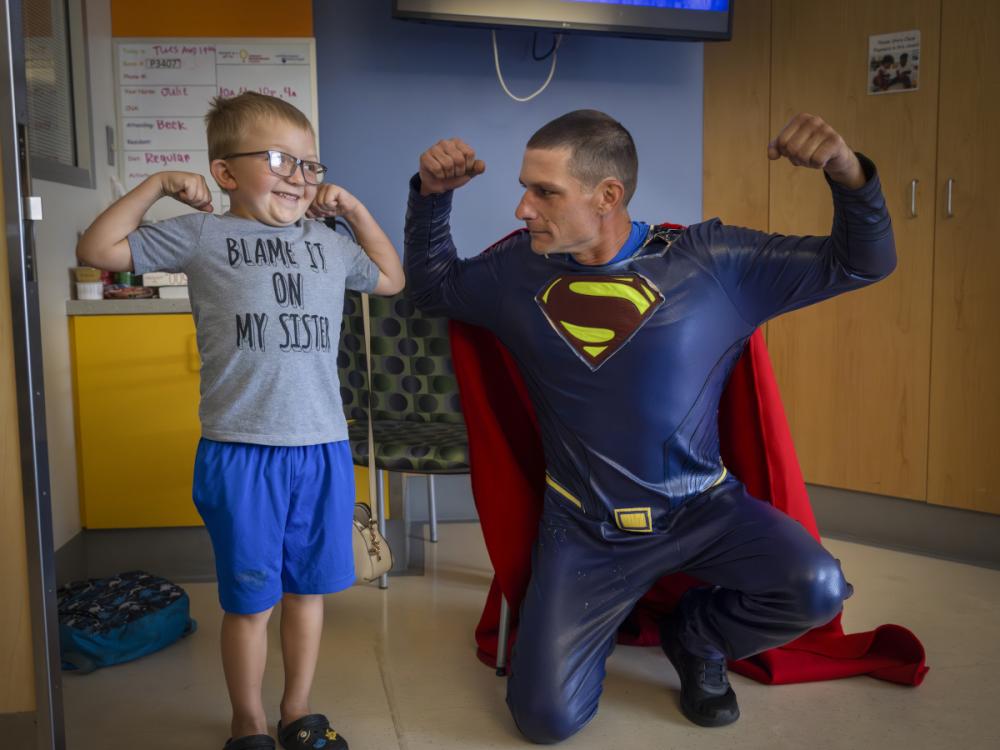 A young boy wearing glasses, a gray T-shirt and blue shorts stands smiling and flexing his arms alongside a man dressed in a Superman costume with a red cape, who is kneeling and flexing his muscles.