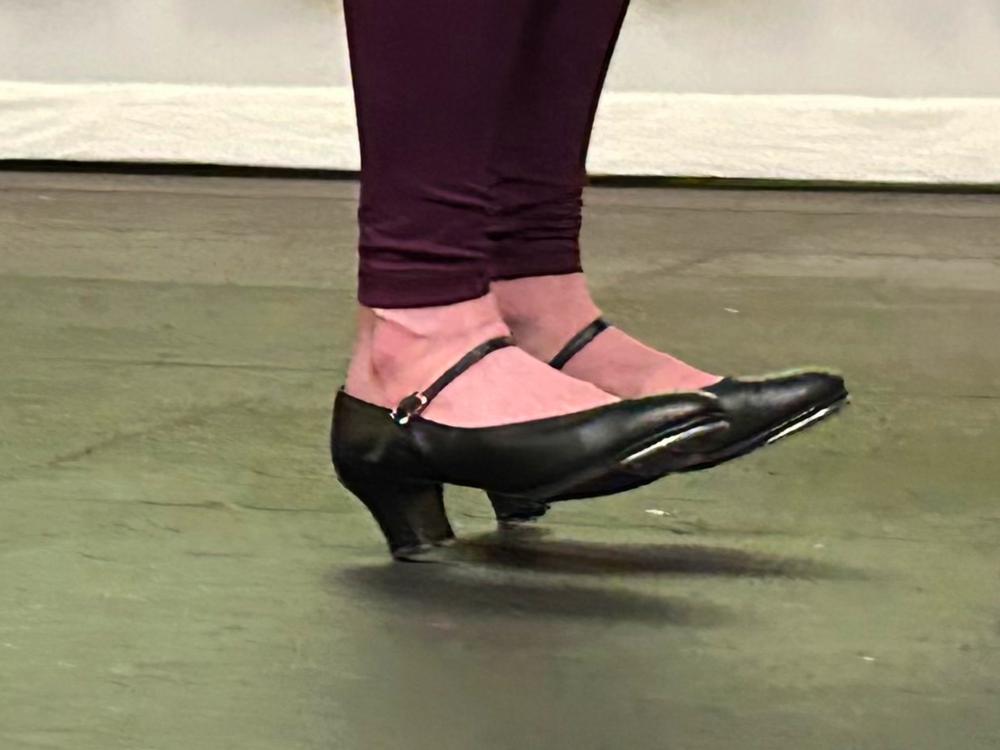 A close-up photo of a dancer's feet mid-air, wearing black character tap shoes with a low heel and ankle straps.