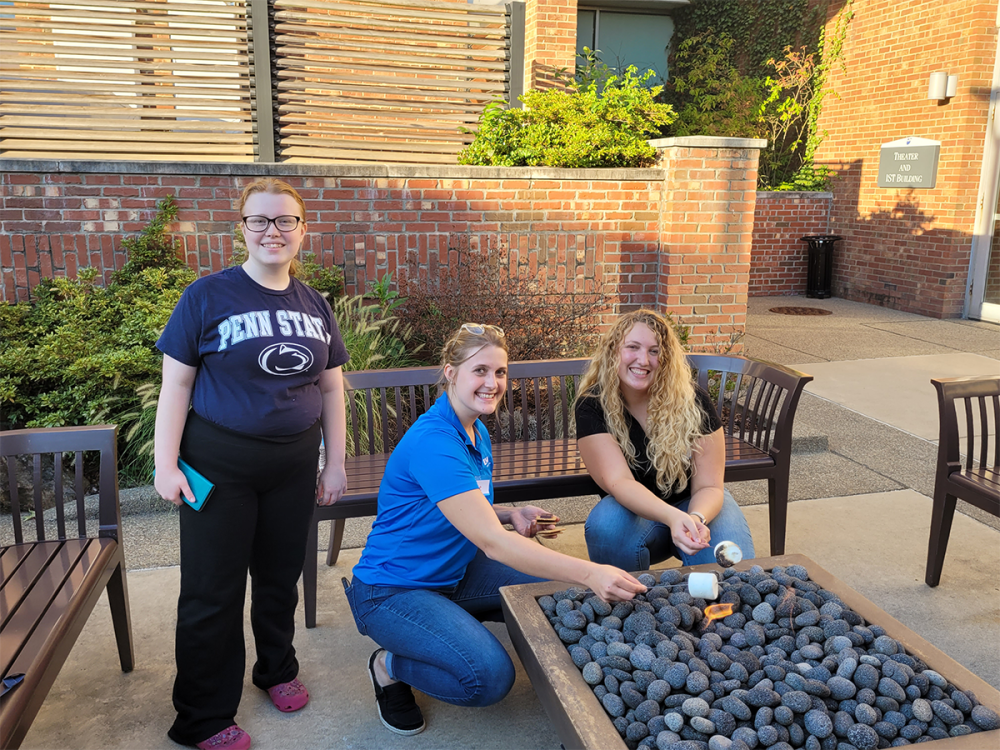 Three women roast marshmallows beside a campus fire pit.