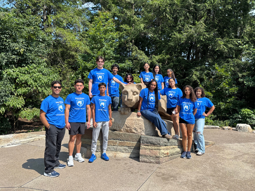 college students in blue t-shirts gather around a Penn State lion shrine and smile on a sunny summer day.