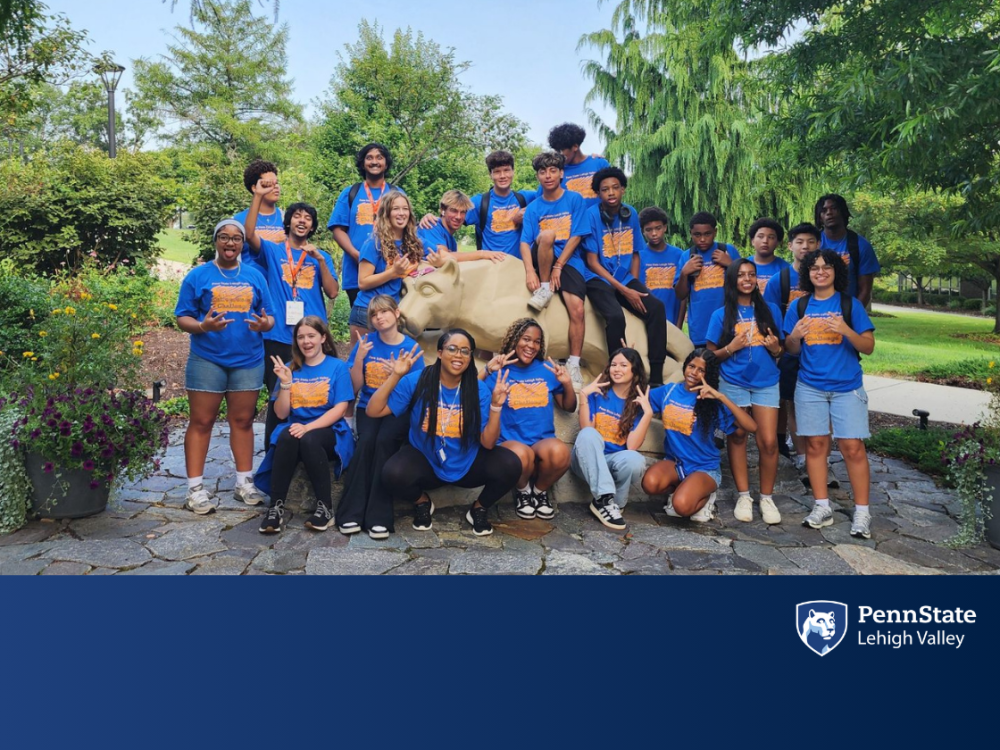 A group of high school students in blue t-shirts smile in front of the Penn State lion shrine