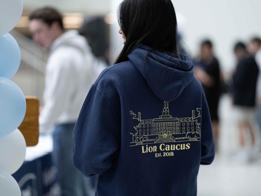 A student wearing a navy blue hoodie with 'Lion Caucus Est. 2018' and an outline of Old Main on the back stands near a table with balloons, while others gather in the background.