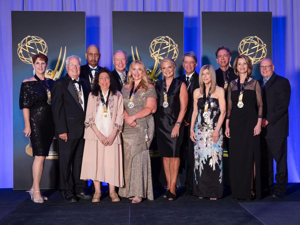 Twelve men and women, dressed in formal attire and wearing medals around their necks, pose in front of a blue and purple backdrop.