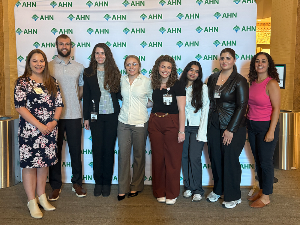 A group of eight people, including Penn State students and mentors, stand together smiling in front of a white Allegheny Health Network step-and-repeat banner. They are dressed in business casual and professional attire, with ID badges visible on several participants.