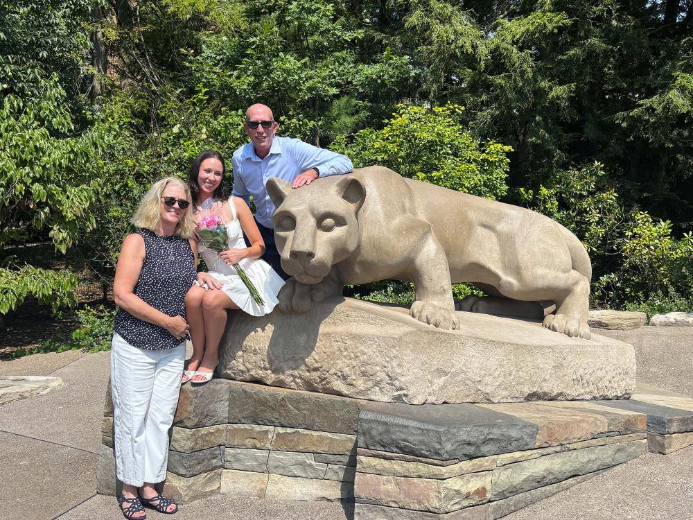 A grad stands in between her parents at the Nittany Lion shrine statue