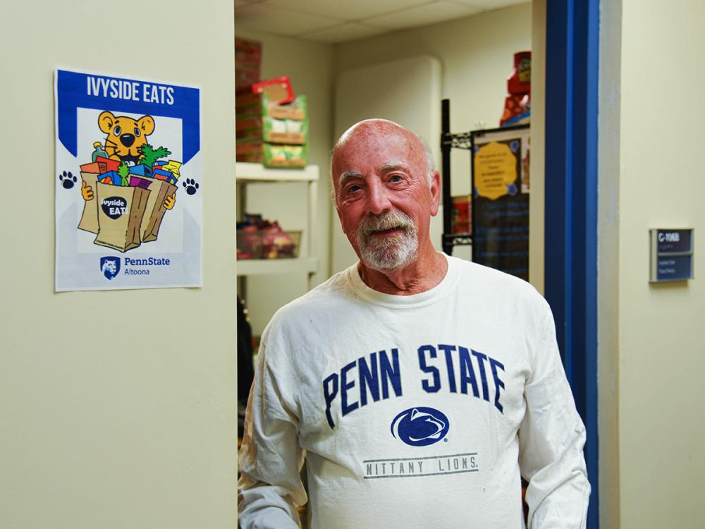 Man in a Penn State shirt stands outside Ivyside Eats food pantry at Penn State Altoona, smiling at the camera.
