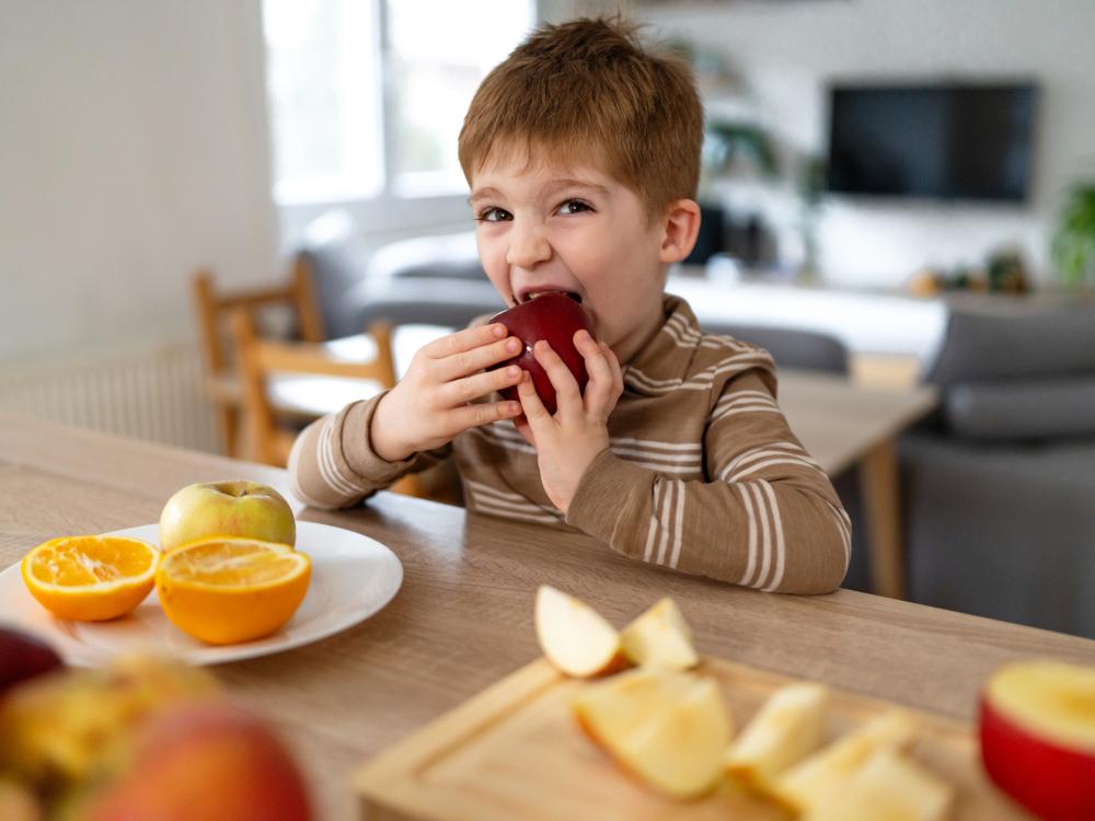A child, sitting at a counter in a kitchen with apple slices and oranges, eats an apple