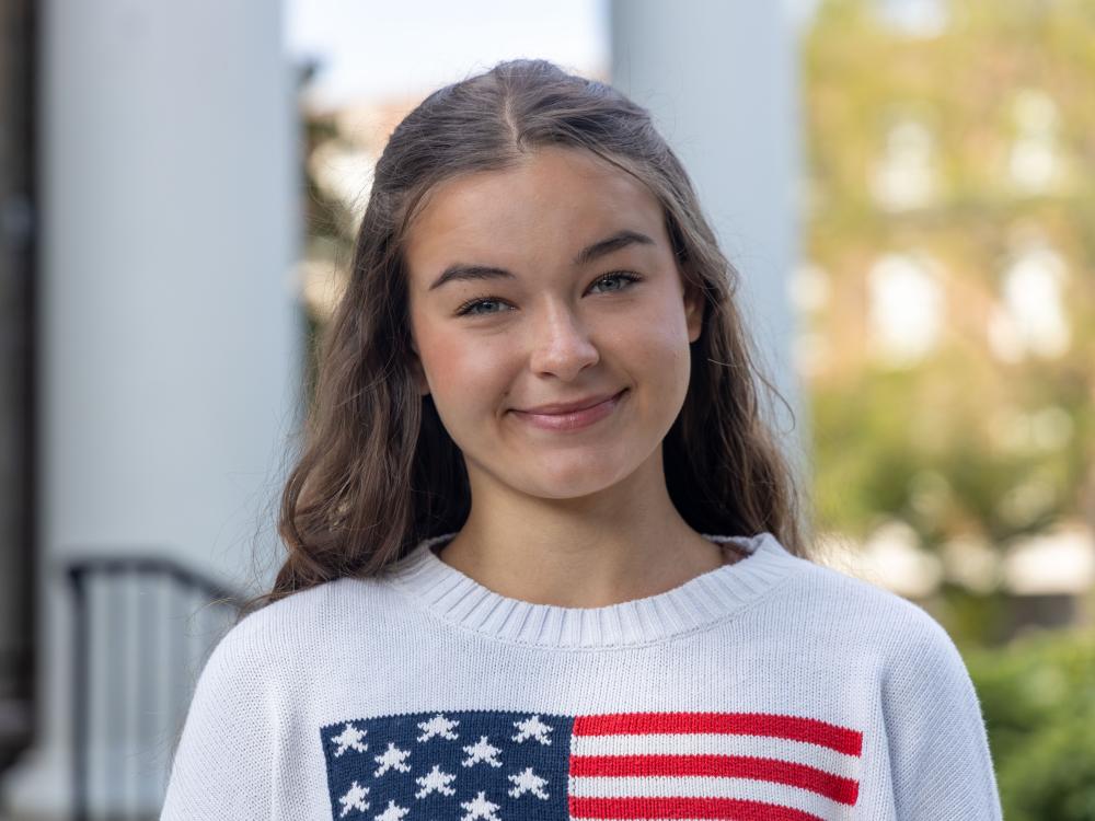 Isabelle Ems wears a white sweater with an American flag on it in front of a backdrop of columns.
