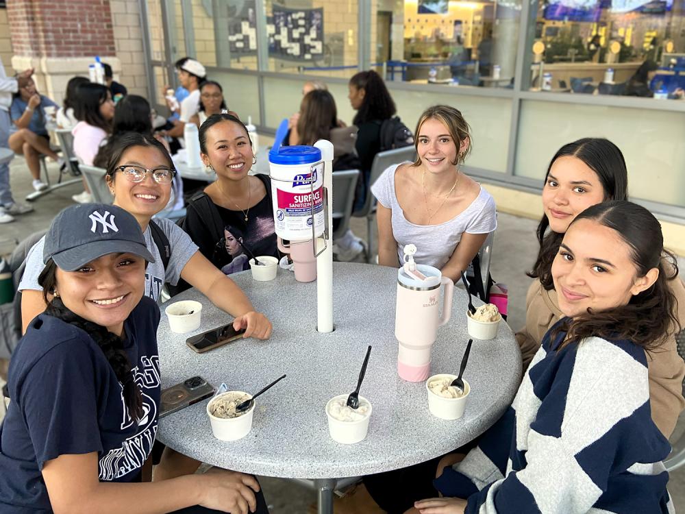 A group of young, female students sit around a circular table with cups of ice cream in front of them.