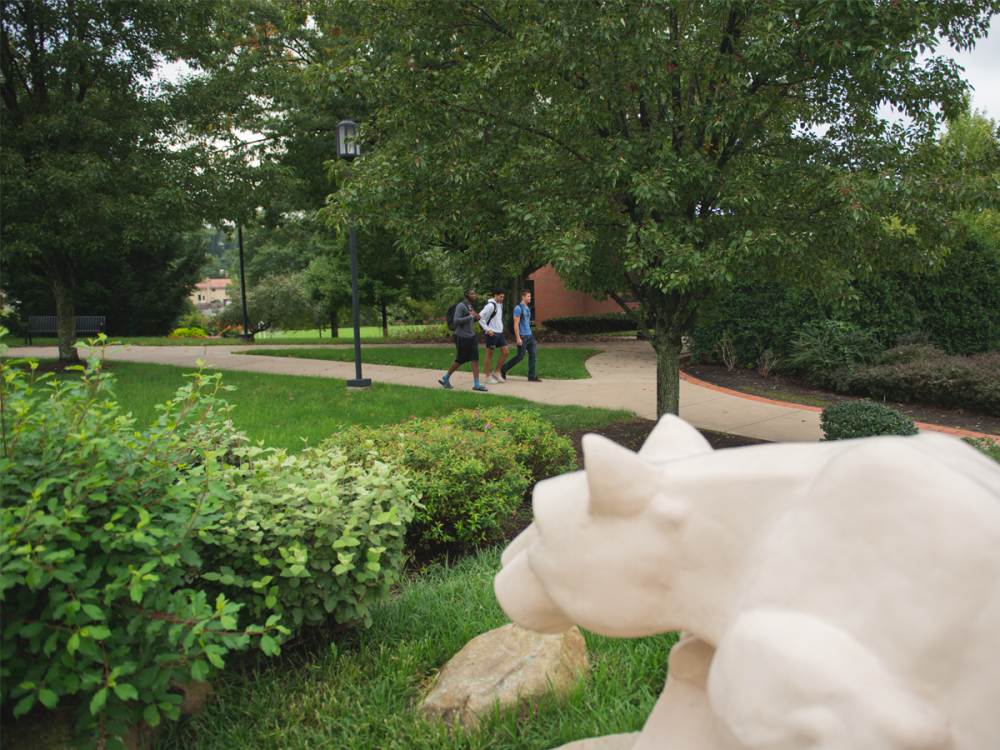 The Nittany Lion shrine statue in the foreground overlooks a tree-lined campus walkway, where three students with backpacks walk together on the path at Penn State Greater Allegheny.