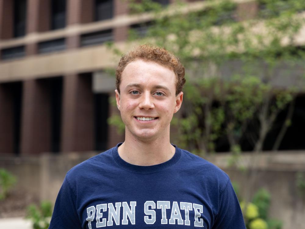 Zachary Graaf stands on a lawn between Ford Building and Welch Building wearing a blue Penn State t-shirt.