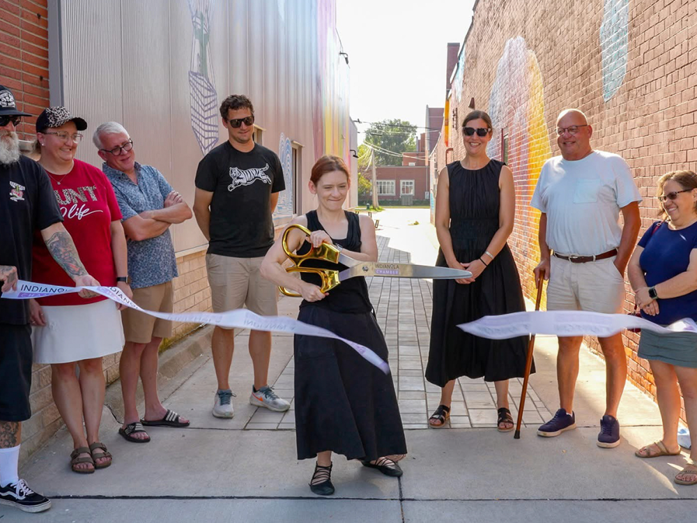 Lori Hepner cuts a ceremonial ribbon with oversized scissors at the entrance of Buxton Alley in Indianola, Iowa, while community members stand smiling on either side of her.