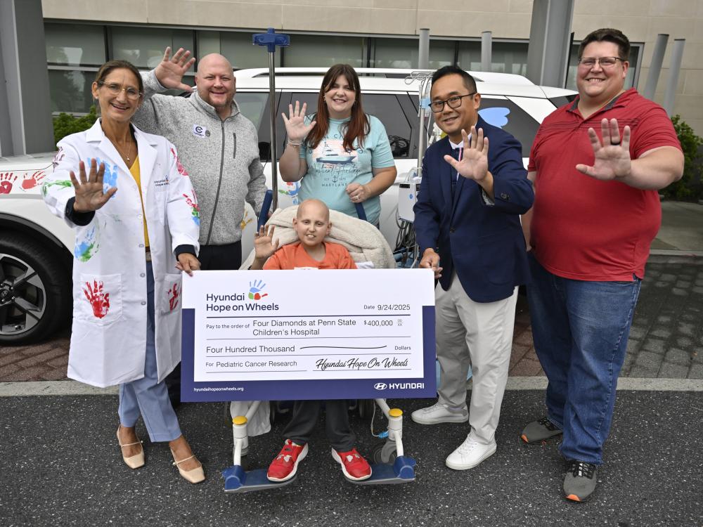 Five people hold up one of their hands toward the camera as they pose for a photo with a check from Hyundai Hope on Wheels for $400,000. A car is in the background, and behind that is Penn State Health Children’s Hospital.