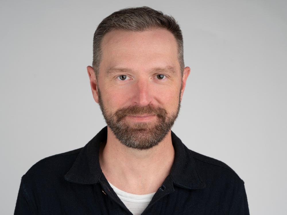 Kirk Goldsberry is a man with short brown hair and a beard, wearing a black collared shirt over a white t-shirt, poses against a plain light gray background.