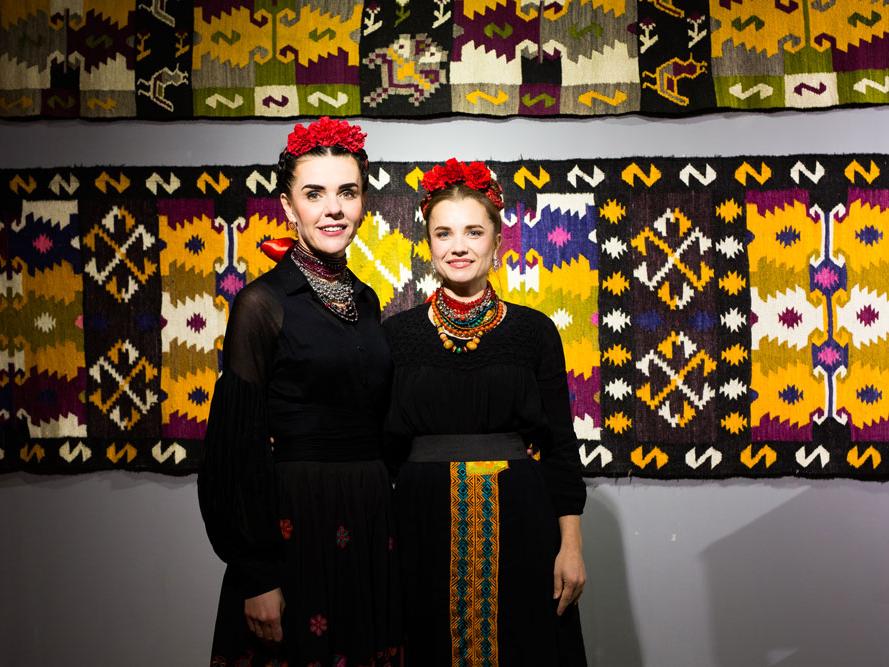 Two women wearing traditional dress stand in front of a mosaic background.