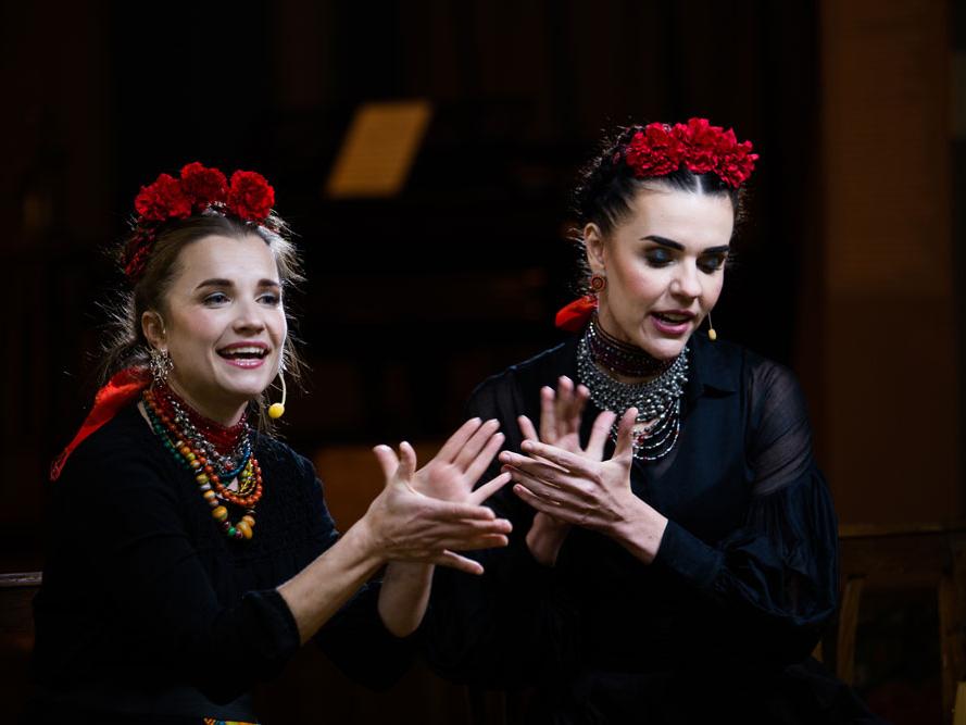 Two women wearing traditional floral headdress sing in unison.