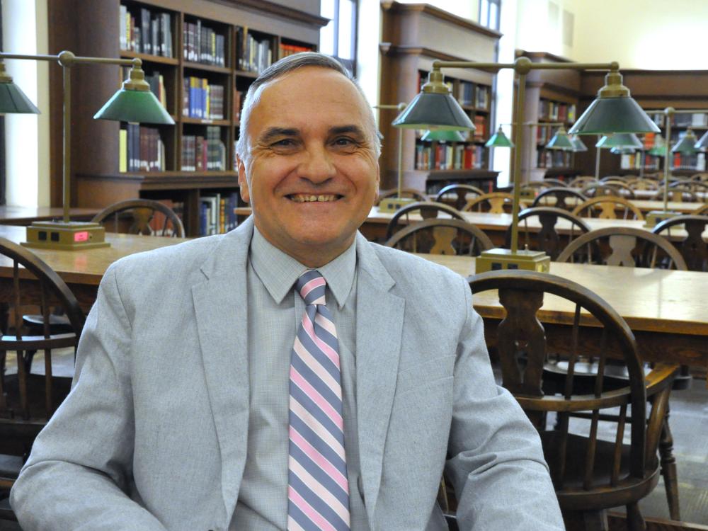 Frantisek Marko smiles at the camera, wearing a gray suit with a grey and pink striped tie. Behind him is a library room with desks and books.