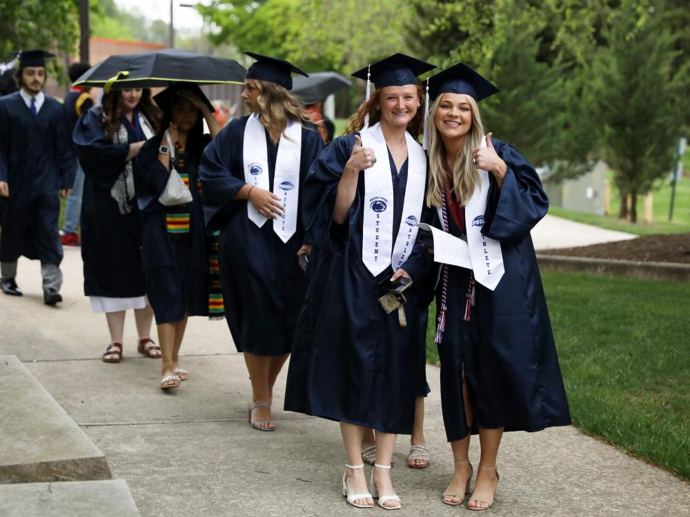 Two students in their cap and gowns at commencement in 2024. the weather is gloomy and one student is holding an umbrella in the background
