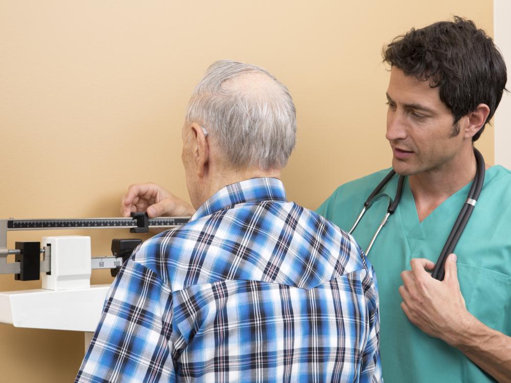 An older person, seen from the back, is standing on a scale while a medical professional weighs them