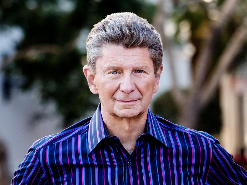 Man with thick graying hair wearing an open collared shirt with black and purple stripes.