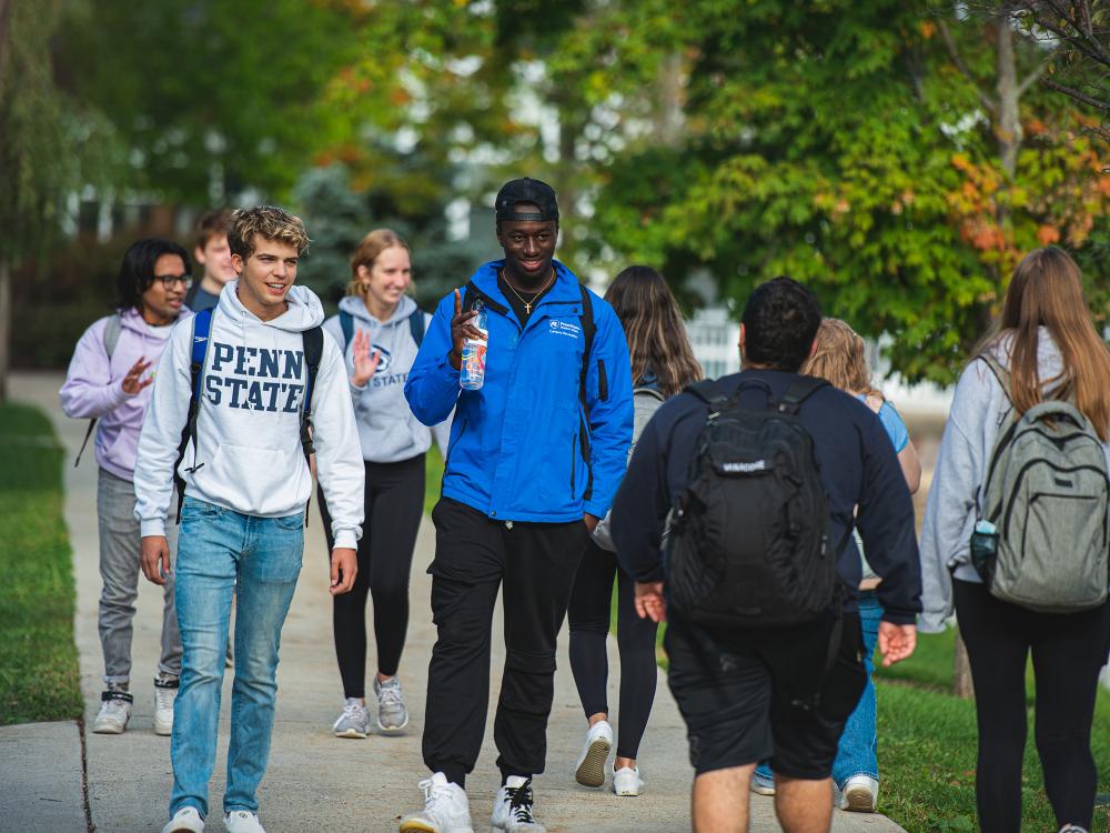 Students walk together on campus, some wearing Penn State gear, chatting and smiling on a tree-lined sidewalk.