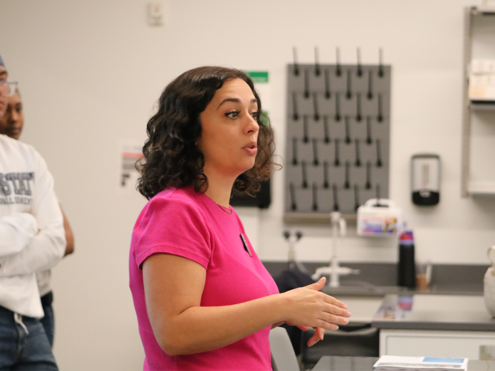 Andrea Stevens, wearing a bright pink shirt, speaks to students during a lab session at Penn State Greater Allegheny. Students stand in the background listening as she gestures with her hands.