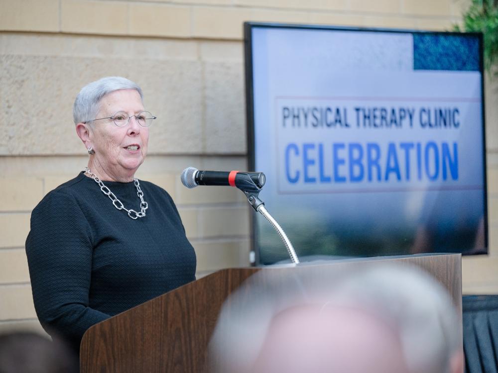 Penn College President Emeritus Davie Jane Gilmour speaks at the dedication ceremony for the college's new Physical Therapy Clinic. A screen next to hear says "Physical Therapy Clinic Celebration."