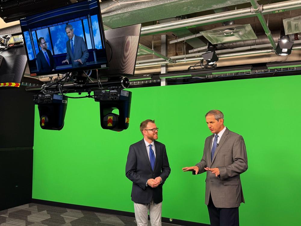 Two men wearing suits talking in front of a green screen on a television broadcast set