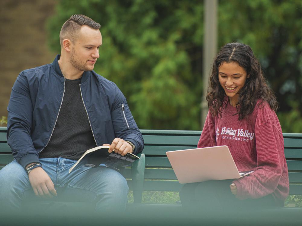 Two students sitting on a bench one has a laptop on their lap