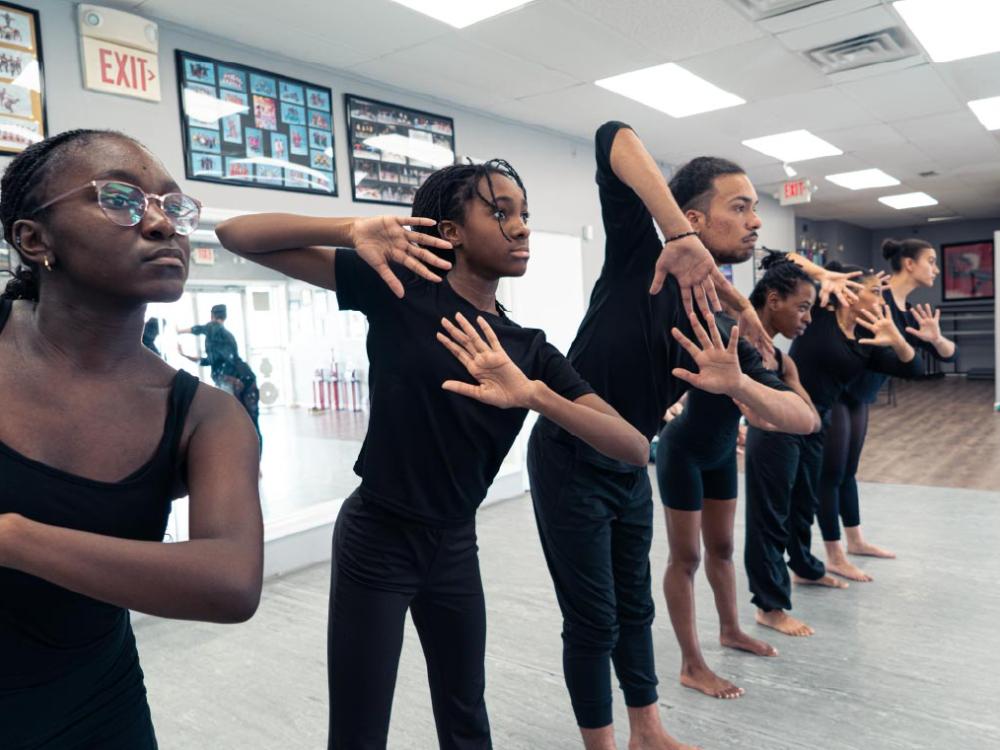 Young Black dancers stand in a row and gesture in unison.