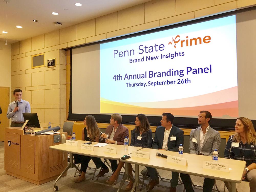 A photo of a student with a microphone looking at six adults, three women and three men, sitting at a table with a graphic that reads "Penn State Prime Brand New Insights" on a screen behind them.