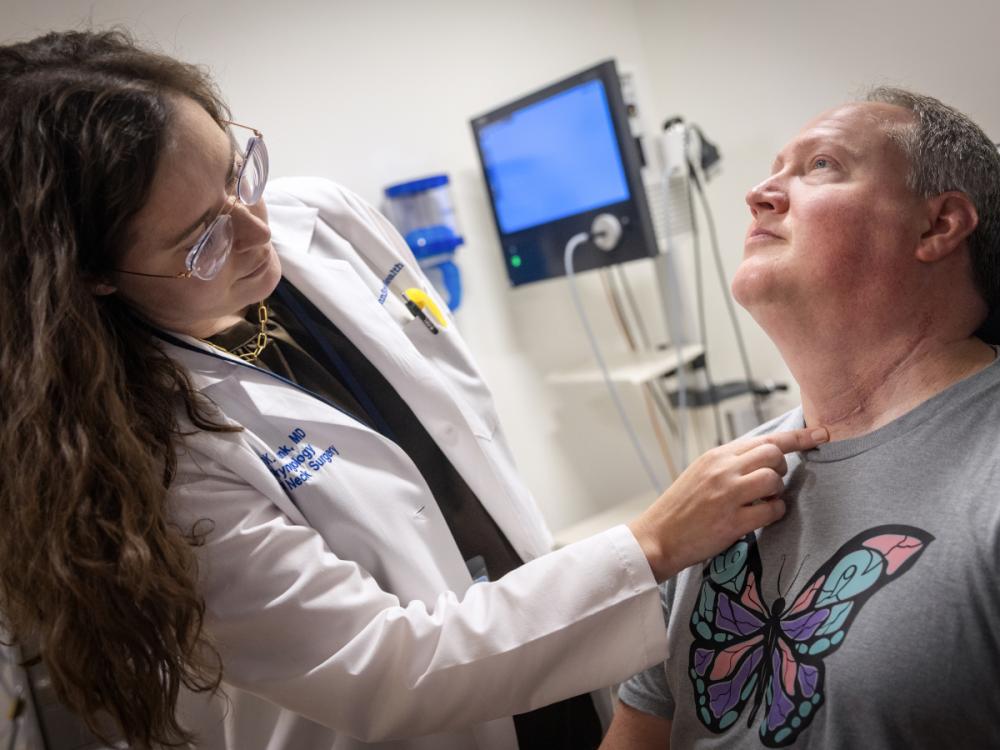 Dr. Emily Funk of Penn State Health Otolaryngology – Head and Neck Surgery points to a scar on the neck of a male patient in an exam room. She is wearing a white lab coat and glasses. He is wearing a T-shirt. Behind them is a monitor.