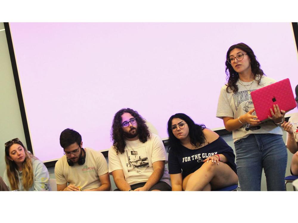 Female student holding laptop addresses a group of students at event