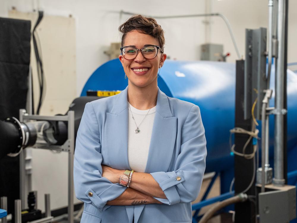 Tamy Guimarães, wearing glasses and a blue jacket, stands in front of a wind tunnel in a lab.
