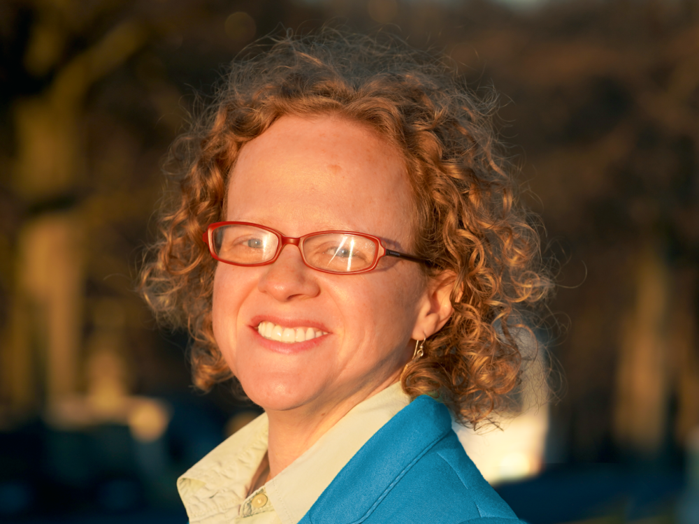 A headshot of a woman in a blue blazer with curly hair and glasses