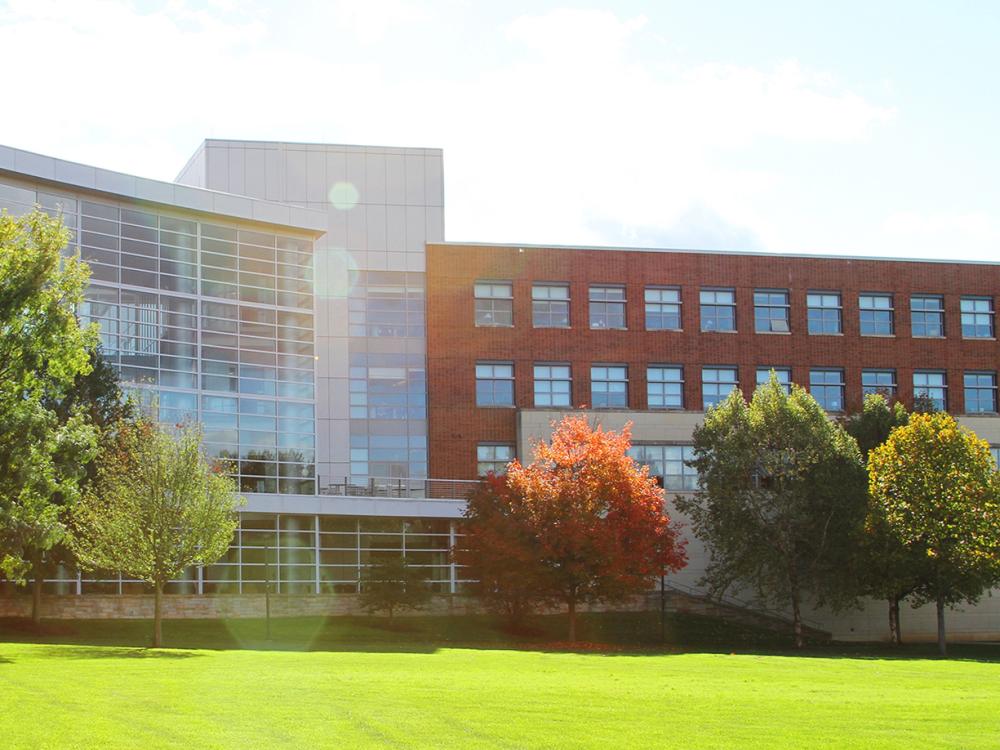 A photo of the Penn State Business Building with the sun shining a tree with orange leaves in front