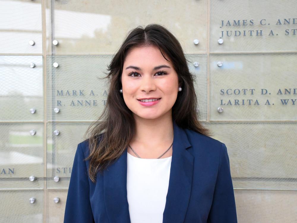 Olivia Cimbora wears a navy blazer and white blouse while standing in front of a glass wall with names etched on it. 