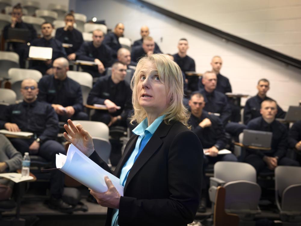 Terry Brosche of Penn State Health stands in front of a group of Pennsylvania State Police cadets who are sitting in an auditorium. She is gesturing, looking up and holding papers. She is wearing a suit jacket and blouse.