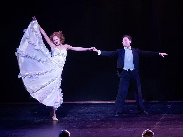 A man and woman dance onstage. The woman is kicking her leg up, flaring the skirt of her white dress.