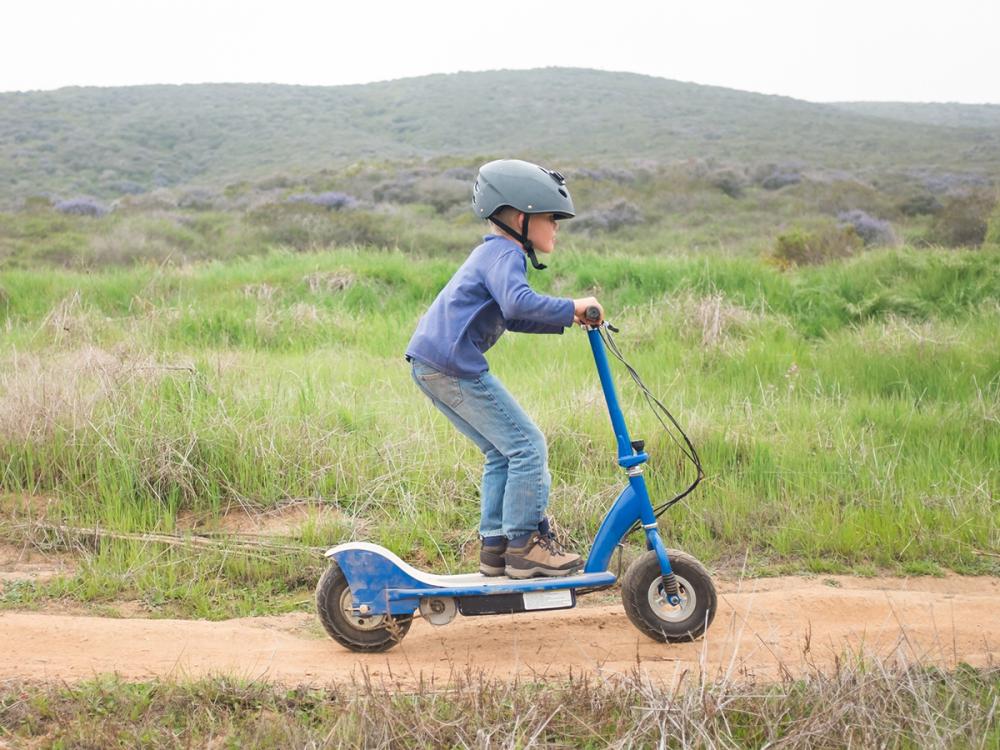 A young boy wearing a helmet rides an electric scooter on a dirt path.