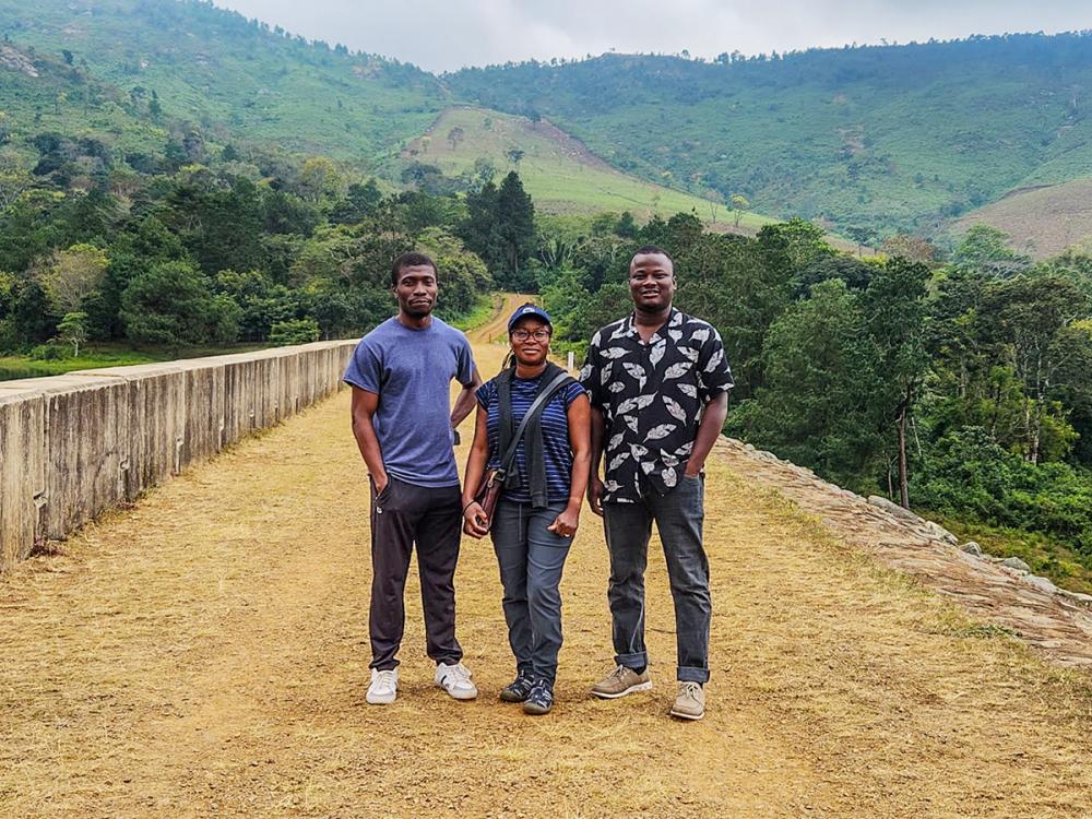 Three people stand on a dirt pathway with lush green hills in the background in Malawi.
