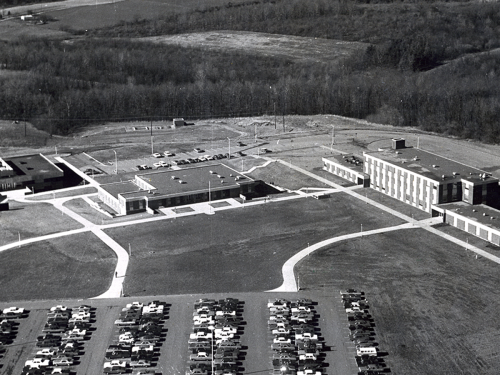 Black-and-white aerial photograph of Penn State Fayette, The Eberly Campus, showing several connected academic buildings, parking lots filled with cars, and open fields and woods in the background.