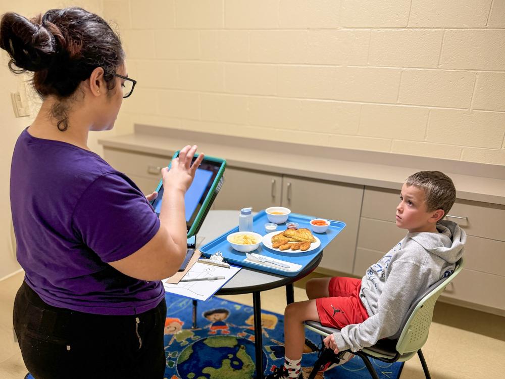A child sits at a table with one of the sample meals used in the study and looks at a research assistant prior to eating.