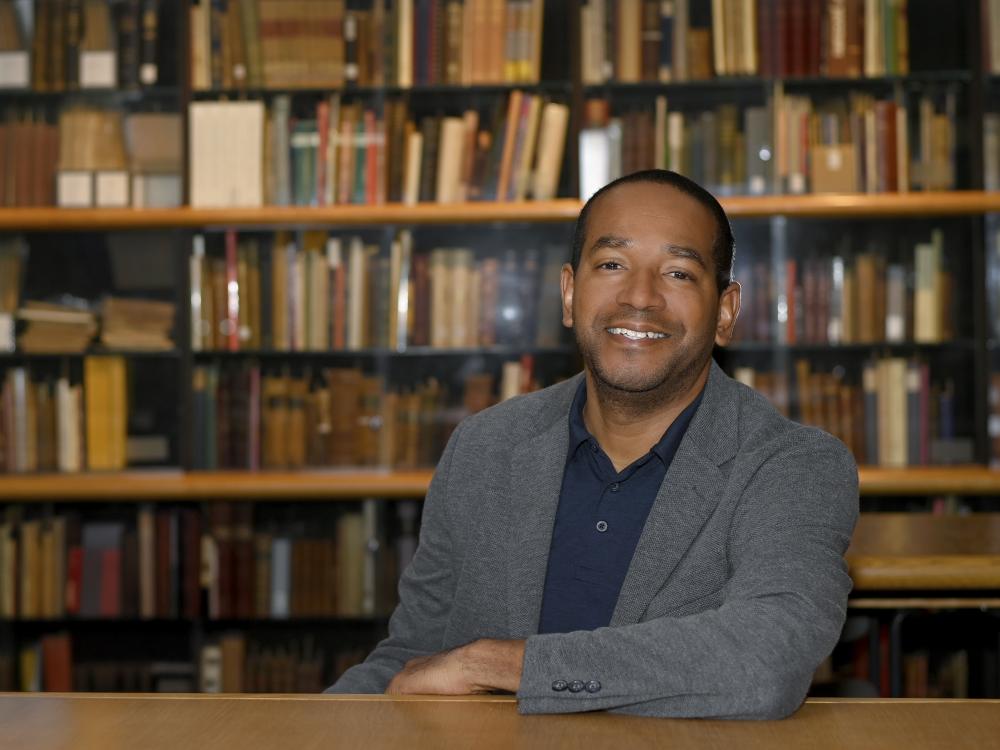 a man in a blue gray suit smile at the camera, sitting at a table in front of a wall of books