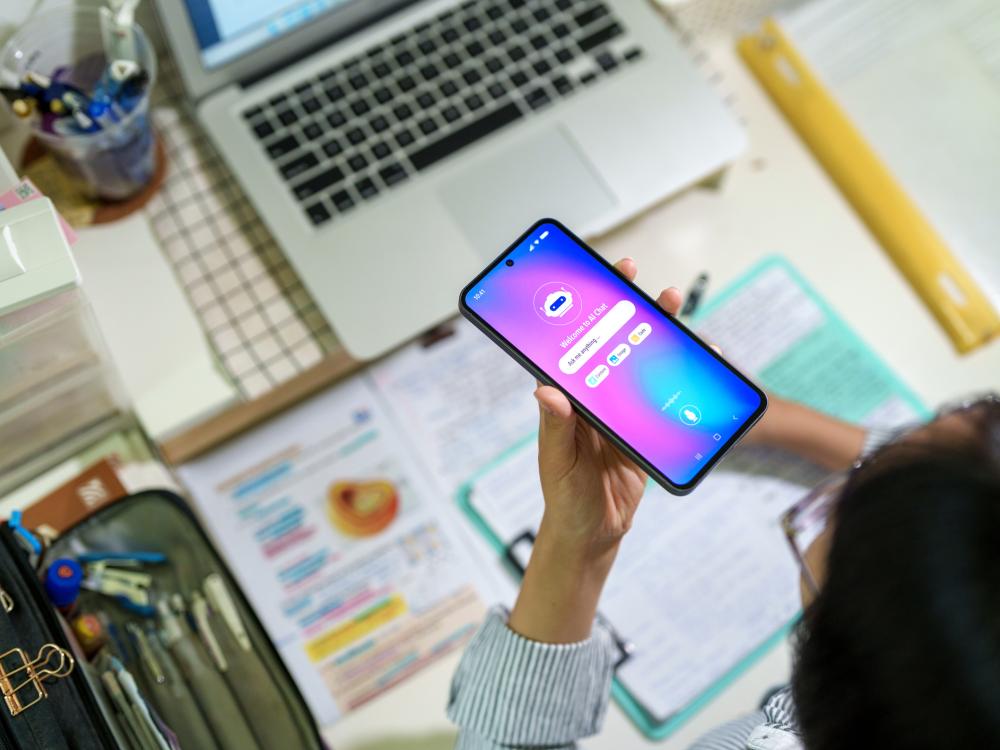 A student interacts with an AI chatbot on a smartphone while at a desk with laptop, notes and stationery.