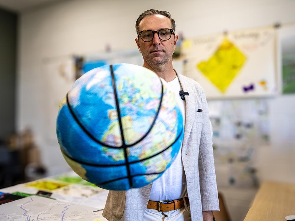 Kirk Goldsberry, wearing glasses and a light blazer, poses with a globe-print basketball in front of maps and charts.